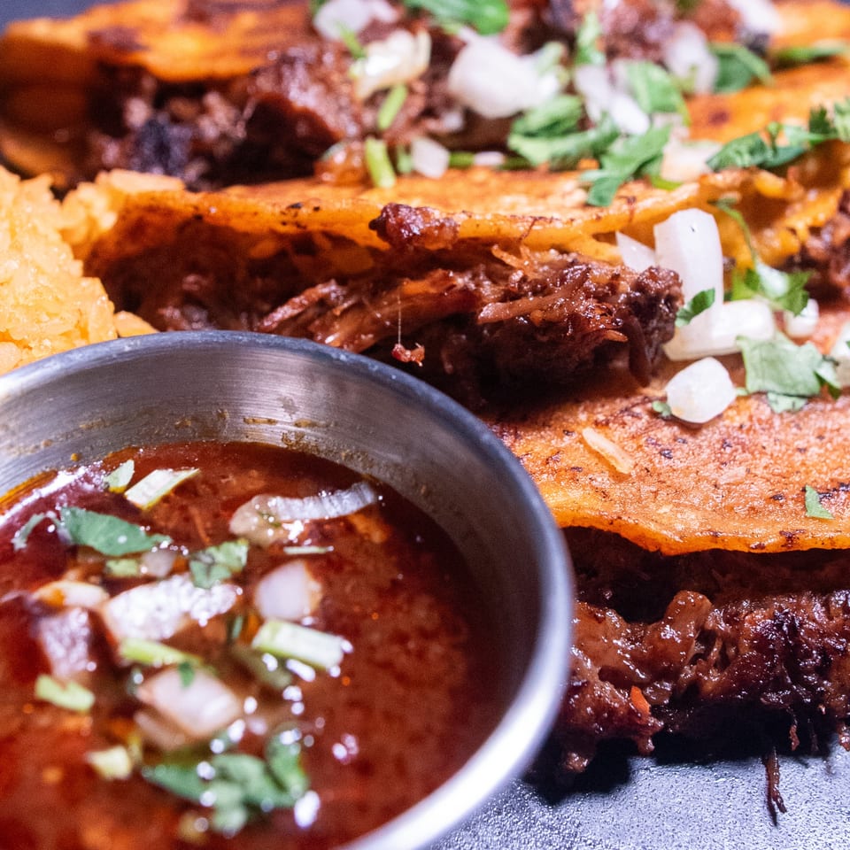 Close-up of birria tacos with rice and a side of consommé, garnished with cilantro and onions.