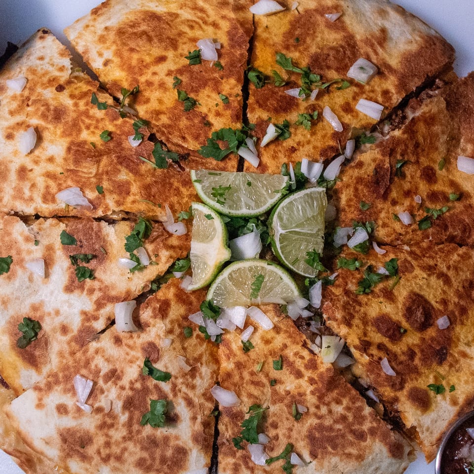 A large, golden-brown quesadilla from Taco Verde, cut into wedges, topped with cilantro, onions, and lime.