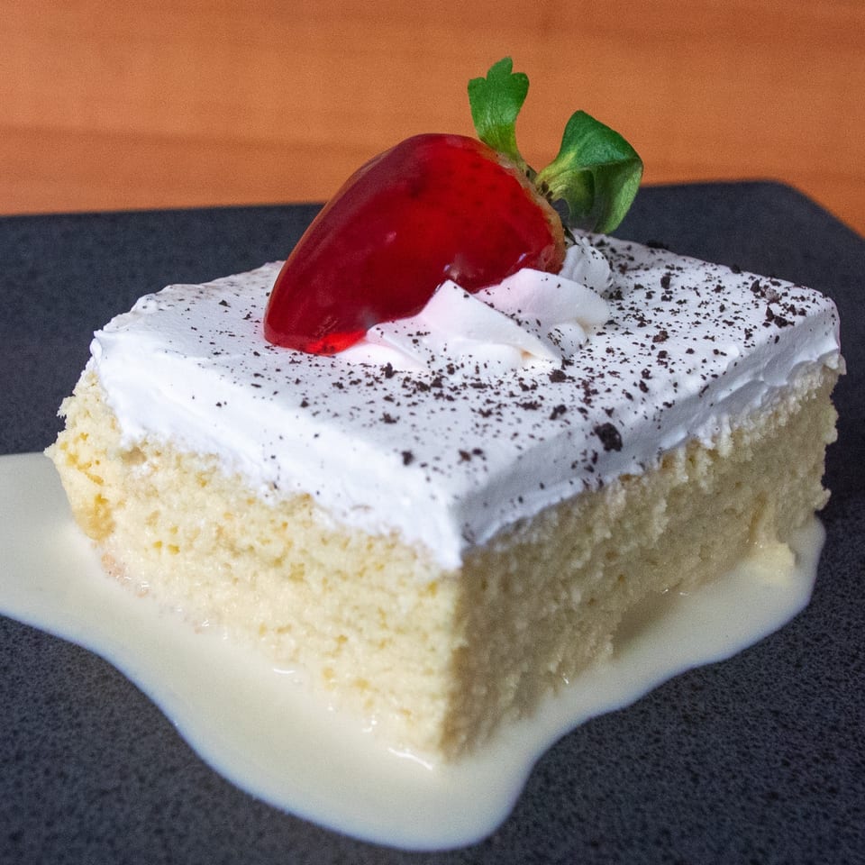 Slice of Tres Leches cake with whipped cream, chocolate sprinkles, and a strawberry on top.